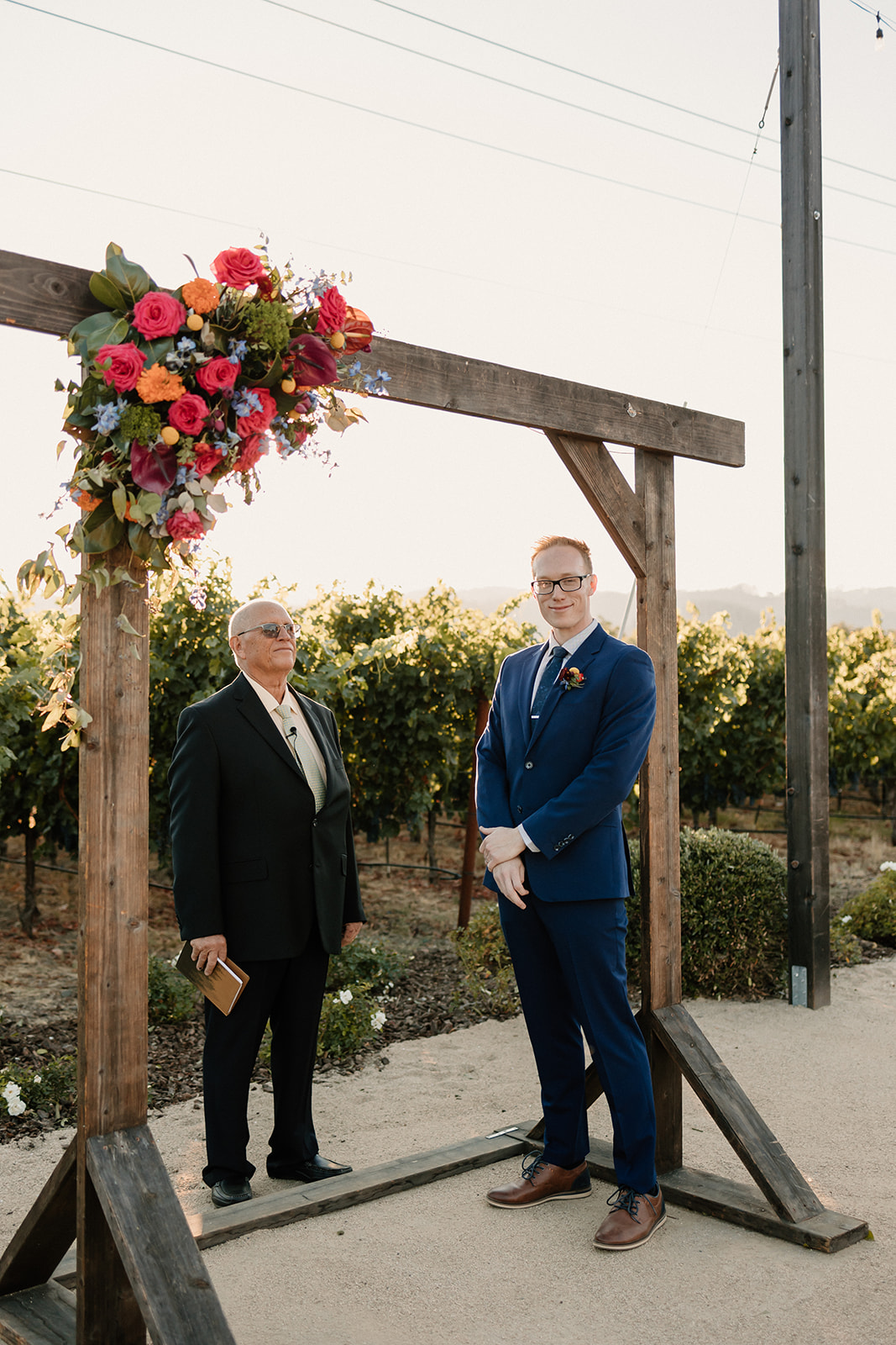 Two men in formal wear standing under a floral arch in a vineyard, one in a black suit and one in a blue suit, smiling gently.