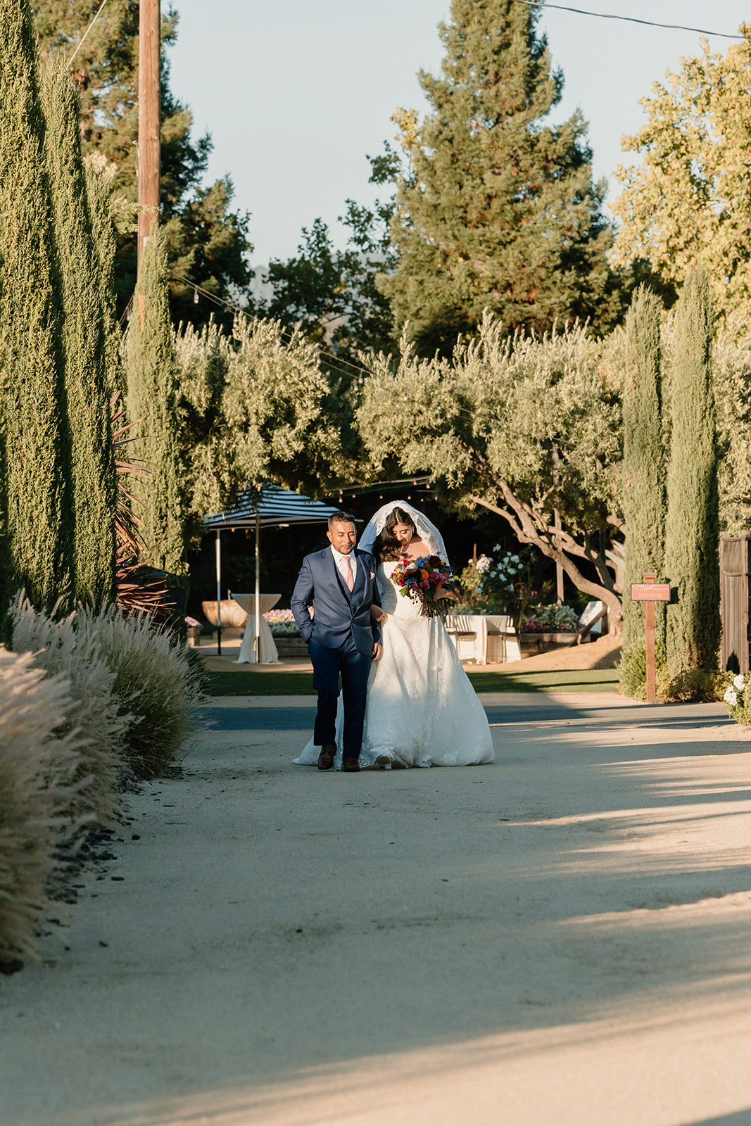 A bride and groom walking hand in hand down a sunlit pathway surrounded by lush greenery and tall trees at Tre Posti wedding venue