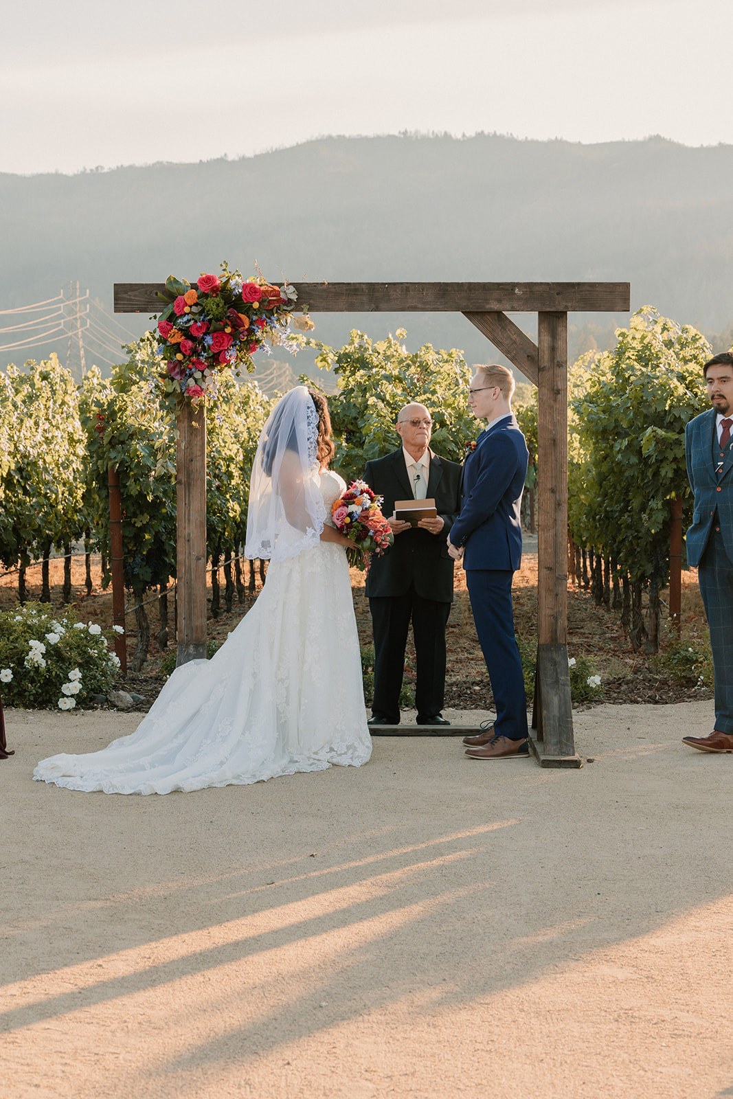 A bride and groom standing at an altar with a clergy member, amidst a vineyard setting during a wedding ceremony at Tre Posti 