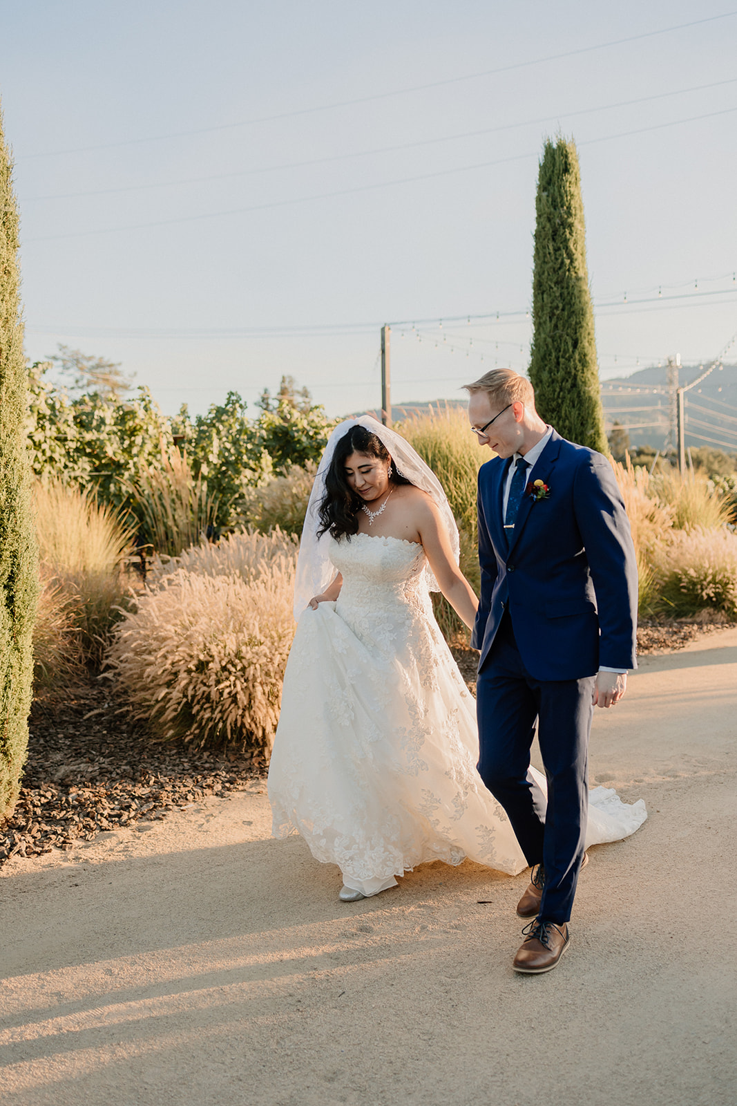 A bride and groom walking hand in hand down a sunlit pathway surrounded by lush greenery and tall trees at Tre Posti wedding venue