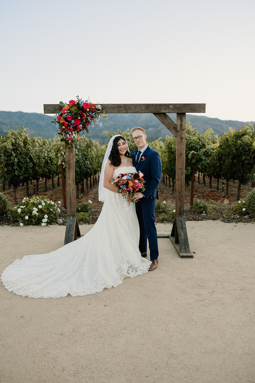 A bride and groom smiling under a wooden arch with colorful flowers, in a vineyard setting.