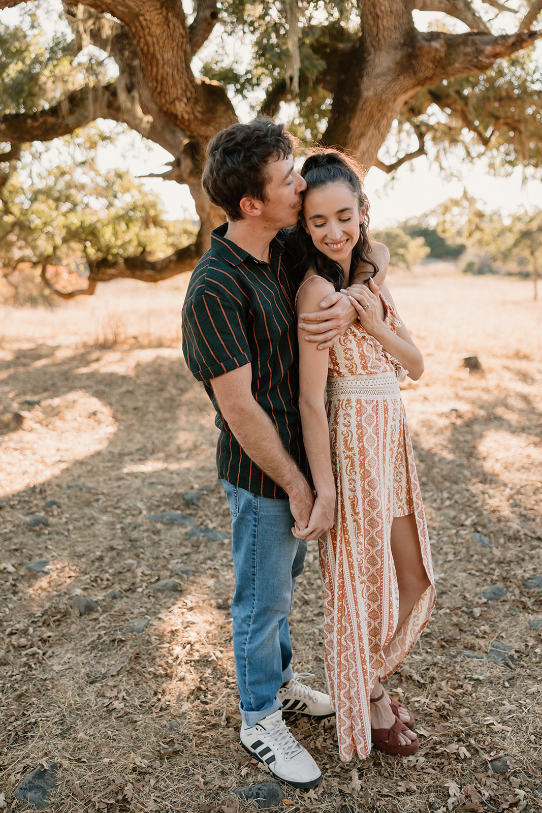 A couple embracing under a tree, exchanging a loving gaze, dressed in casual summer attire for their park engagement photos.