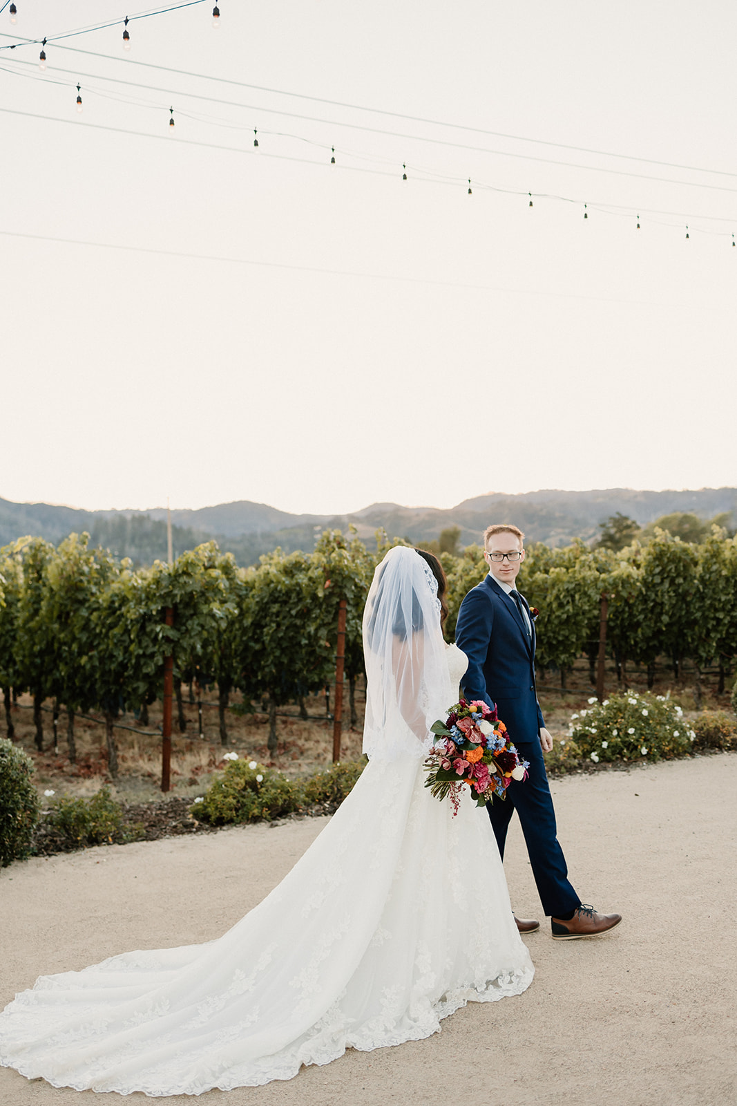 A bride and groom smiling under a wooden arch with colorful flowers, in a vineyard setting at Tre Posti wedding venue
