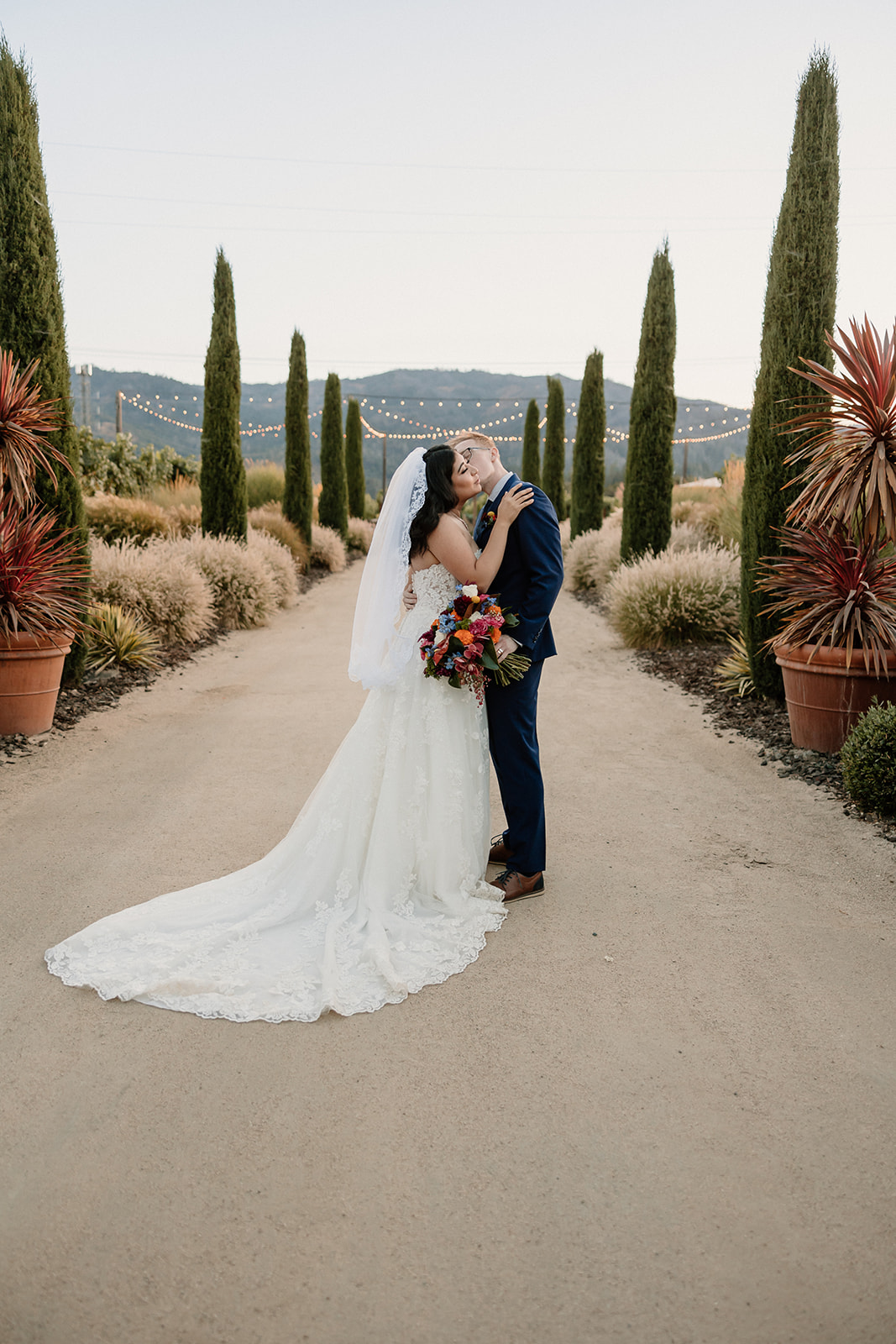 A bride and groom smiling under a wooden arch with colorful flowers, in a vineyard setting at Tre Posti wedding venue