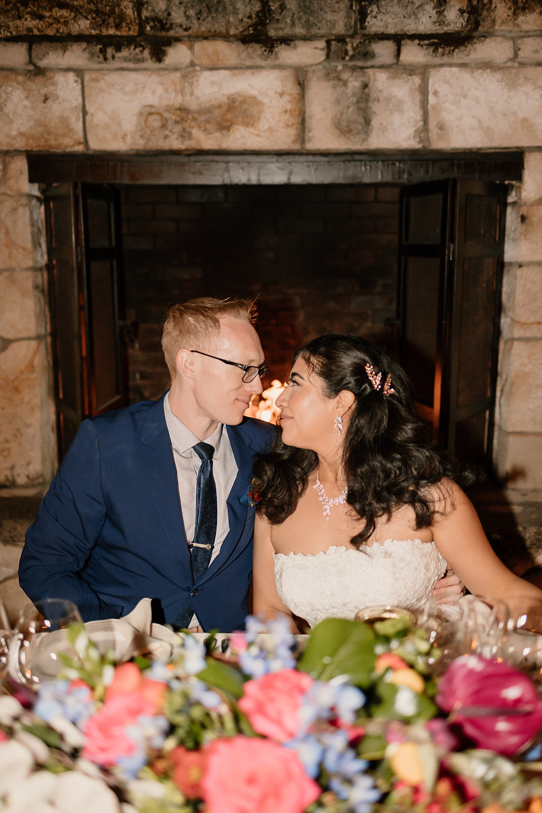 A bride and groom kissing at a banquet table with colorful floral arrangements, in front of a stone fireplace.