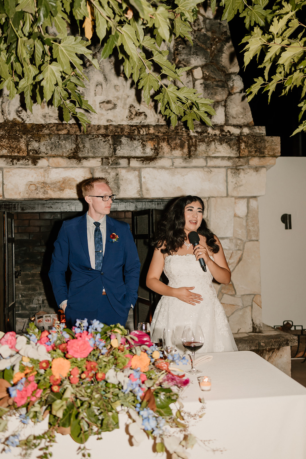 A bride and groom kissing at a banquet table with colorful floral arrangements, in front of a stone fireplace.