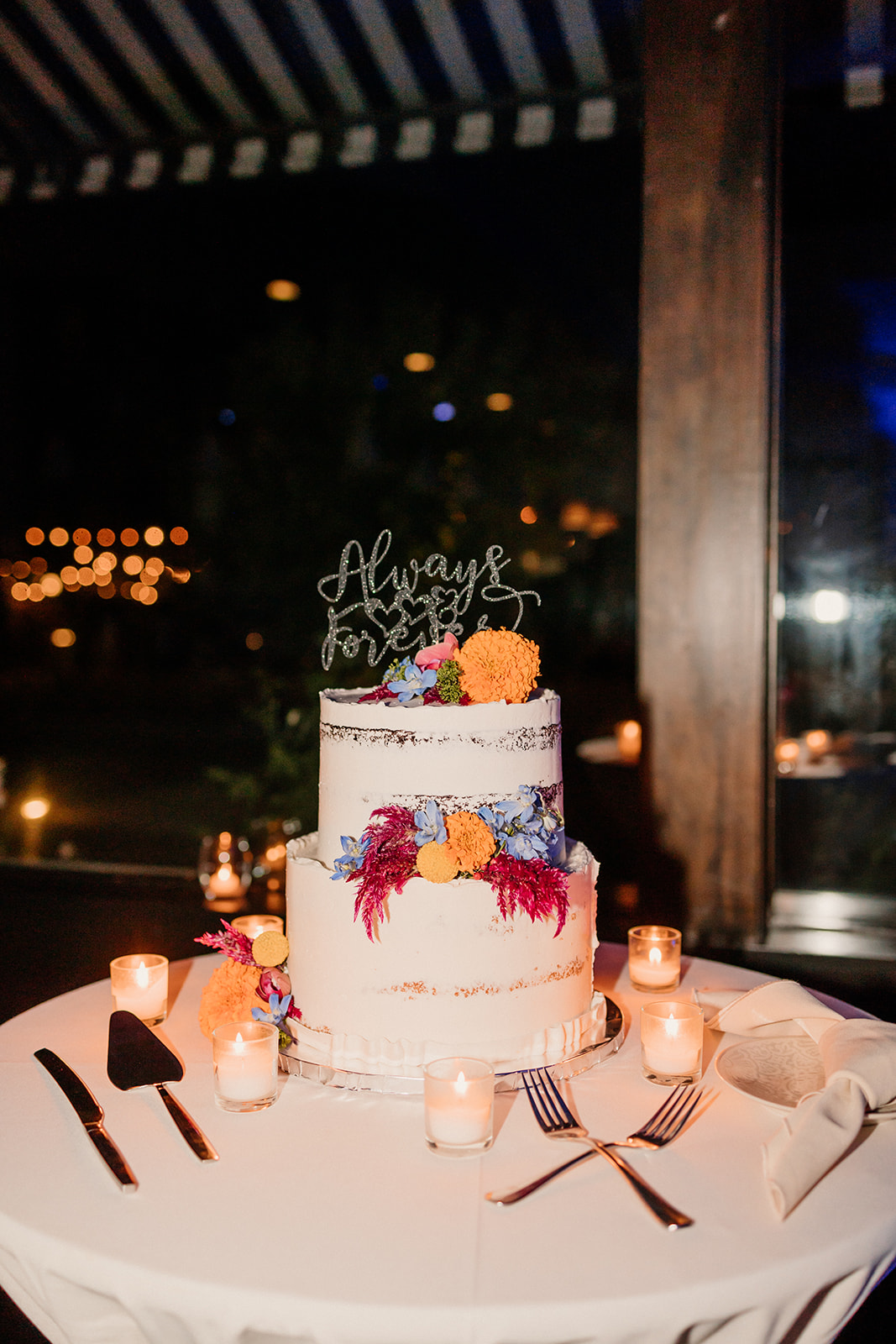 A multi-tiered wedding cake with colorful floral designs on a table, surrounded by lit candles, with a dimly lit background.