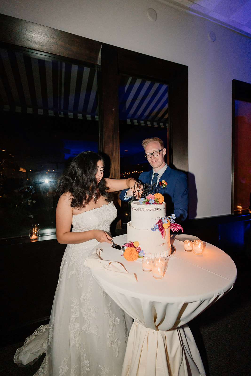 A bride and groom smiling as they cut a wedding cake together at an evening reception.