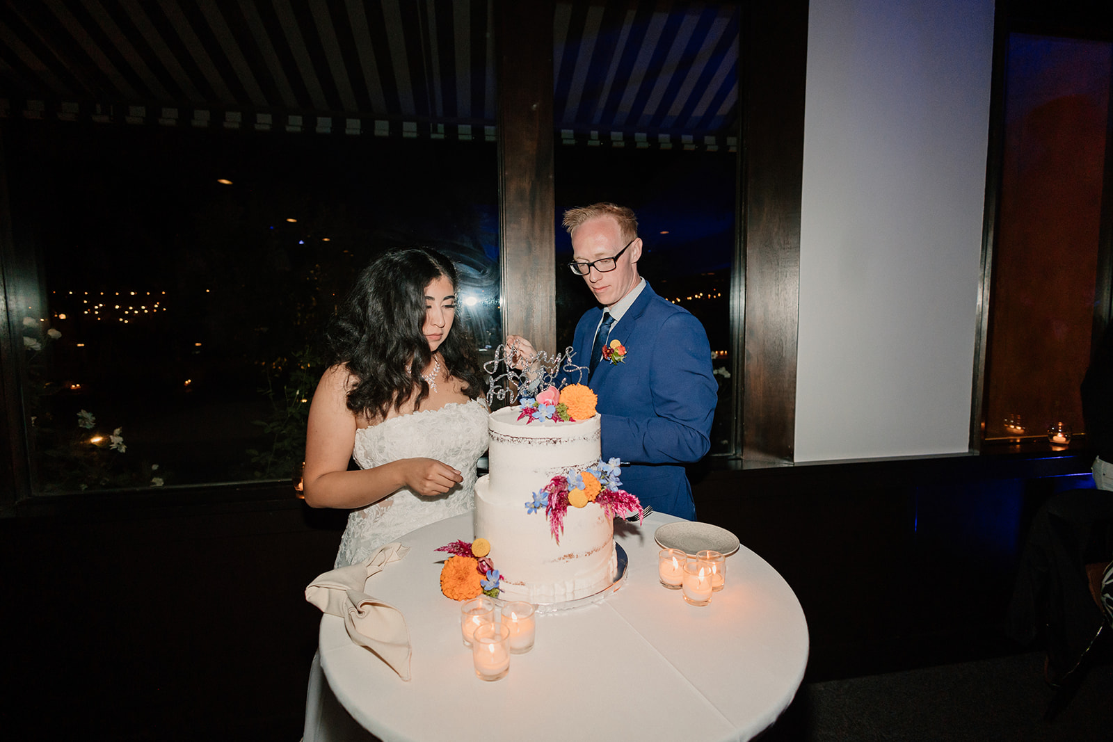 A bride and groom smiling as they cut a wedding cake together at an evening reception.
