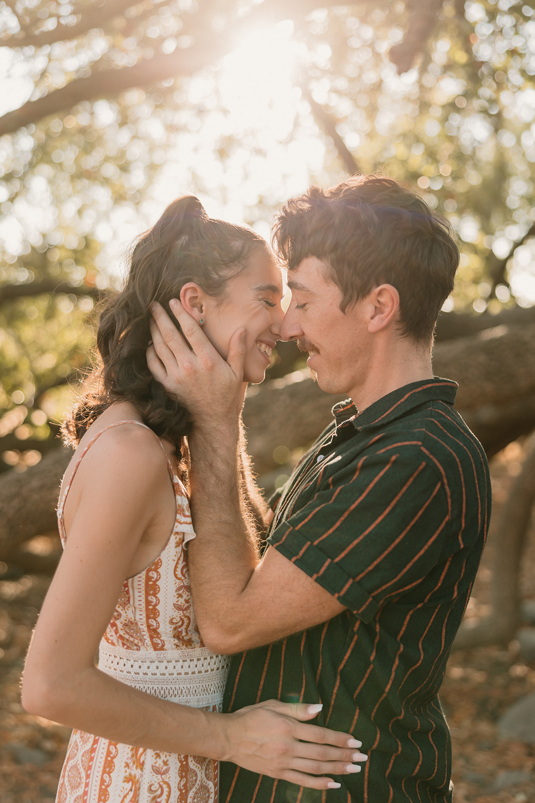 A couple embracing under a tree, exchanging a loving gaze, dressed in casual summer attire for their park engagement photos