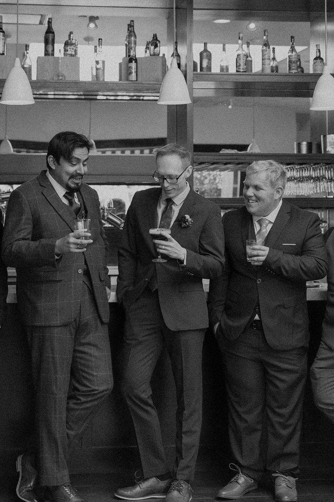 Three men in suits laughing and holding drinks at a bar.
