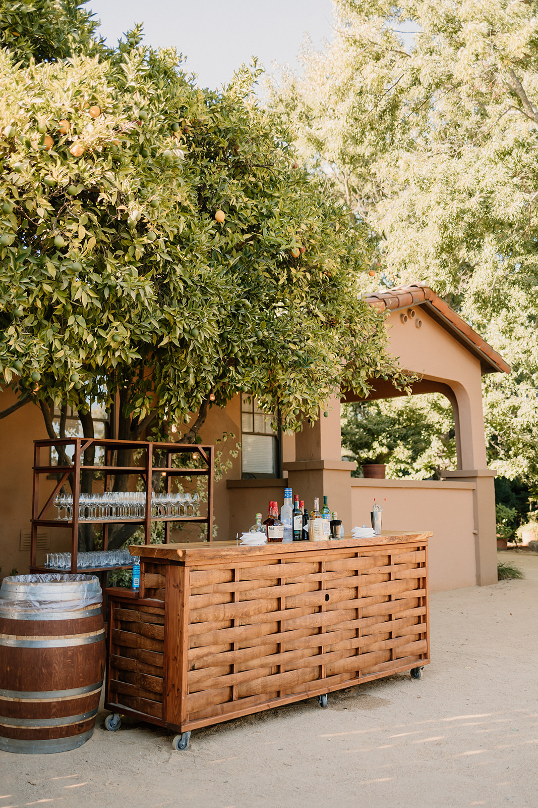Outdoor bar setup under a tree, with a large wooden cart and a variety of bottles on top, next to a building with an arched entrance at Tre Posti Wedding Venue