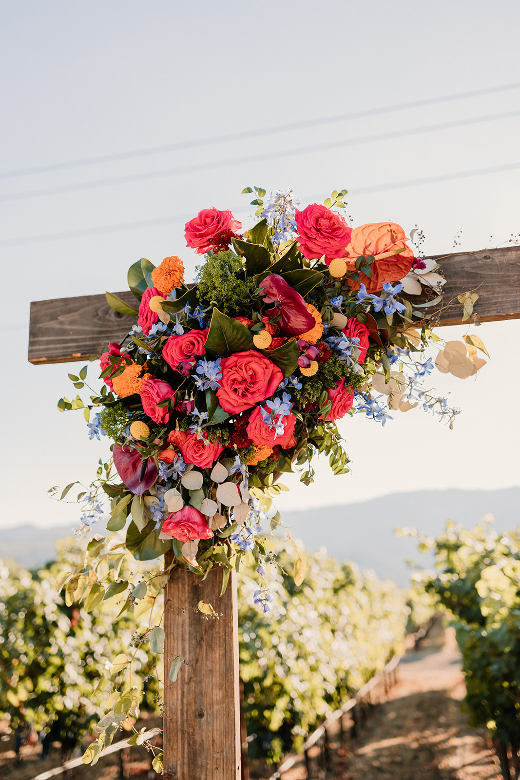 A vibrant floral arrangement on a wooden post, featuring orange, pink, and blue flowers, set against a vineyard backdrop at Tre Posti wedding venue 