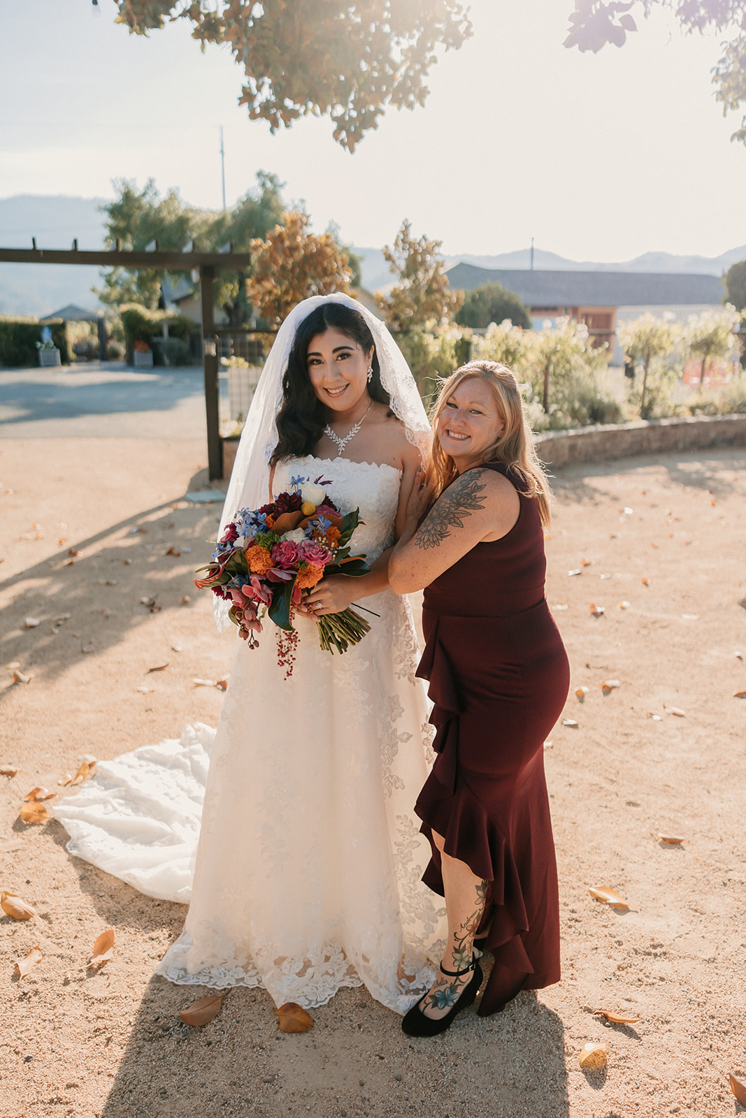 A bride in a white gown and veil holding a bouquet stands smiling with an older woman in a burgundy dress.