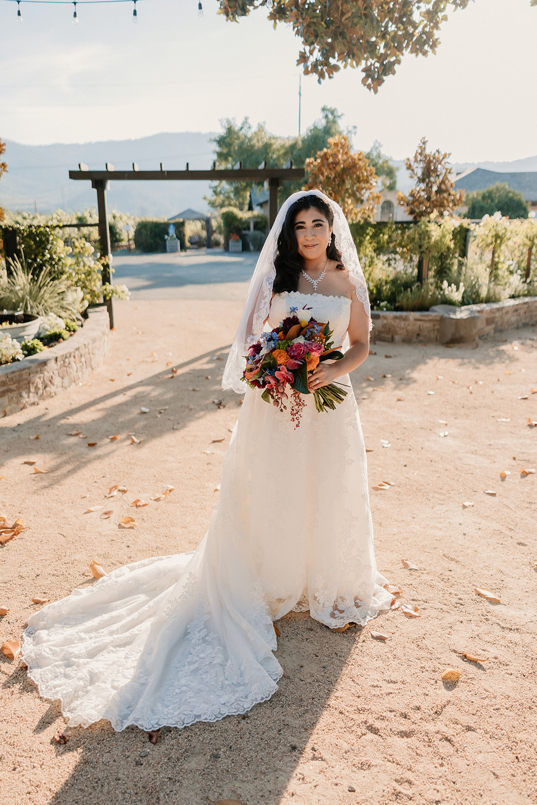 A bride in a white dress and veil holds a bouquet, standing on a sunlit path with fallen leaves at Tre Posti wedding venue 