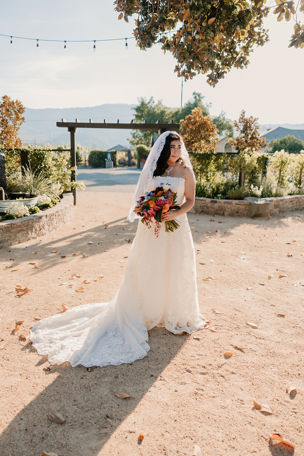 A bride in a white dress and veil holds a bouquet, standing on a sunlit path with fallen leaves at Tre Posti wedding venue 