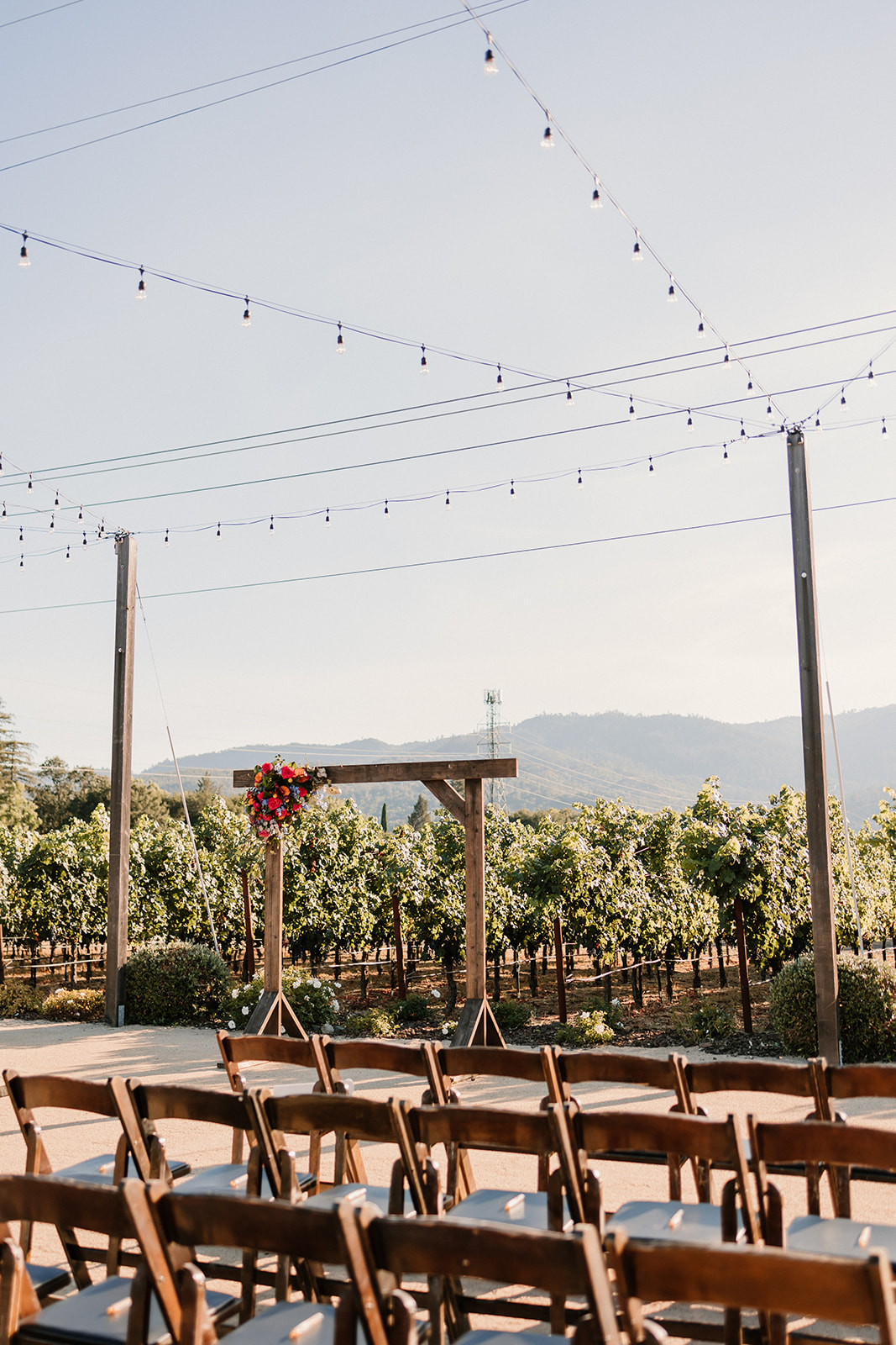 Wooden chairs arranged in rows at an outdoor venue, with a vineyard in the background and string lights above at Tre Posti wedding venue