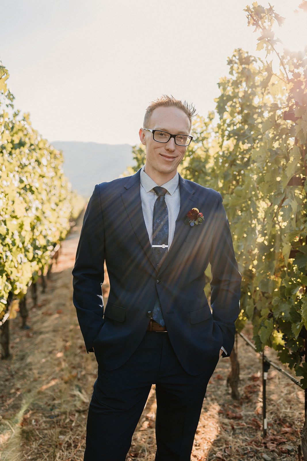 A man in a suit and glasses stands confidently in a vineyard, with sunlight filtering through the grapevines.