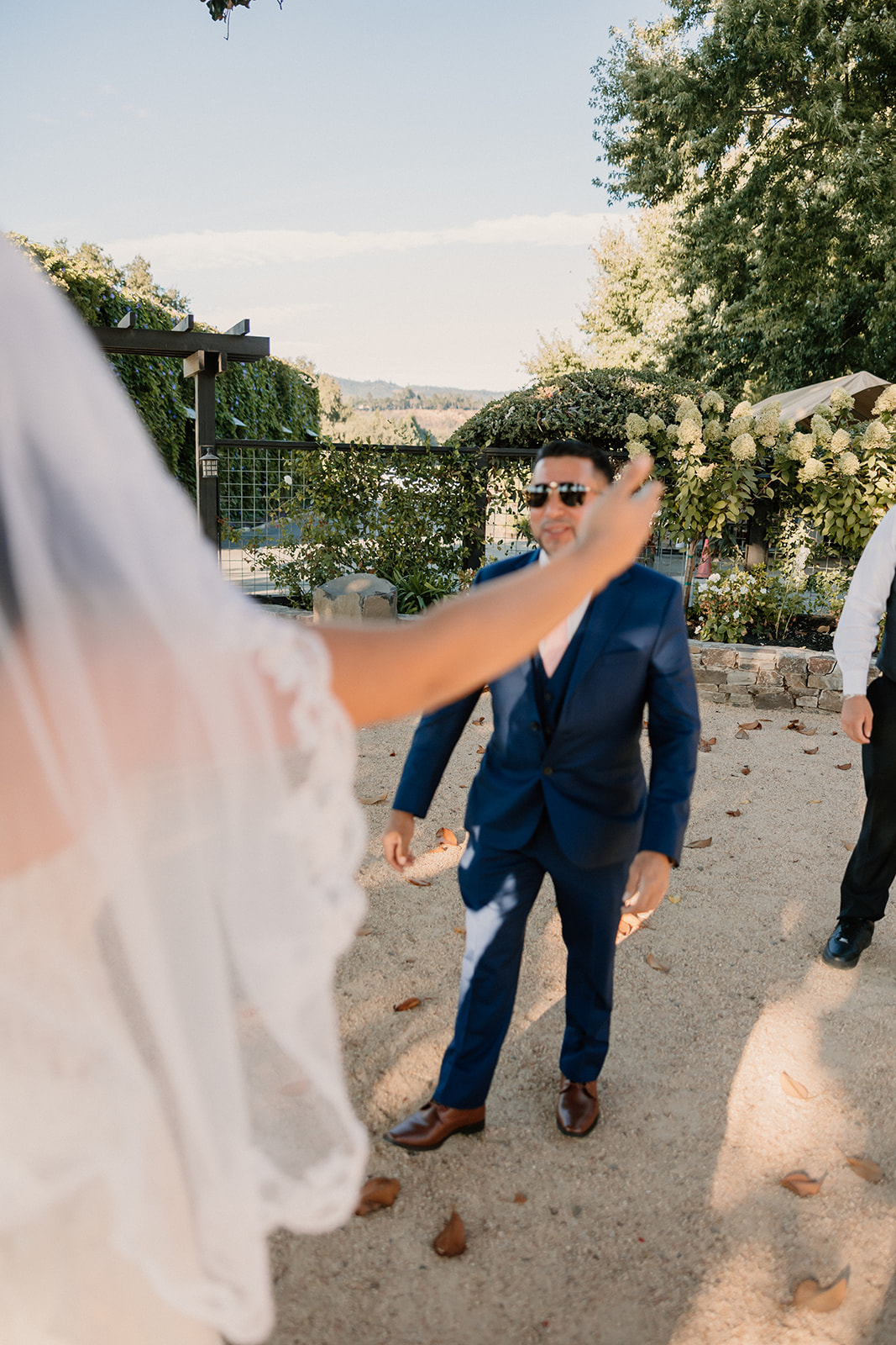 Bride in a lace dress and veil embracing a family member in a blue suit and sunglasses outdoors on a sunny day at Tre Posti Wedding Venue