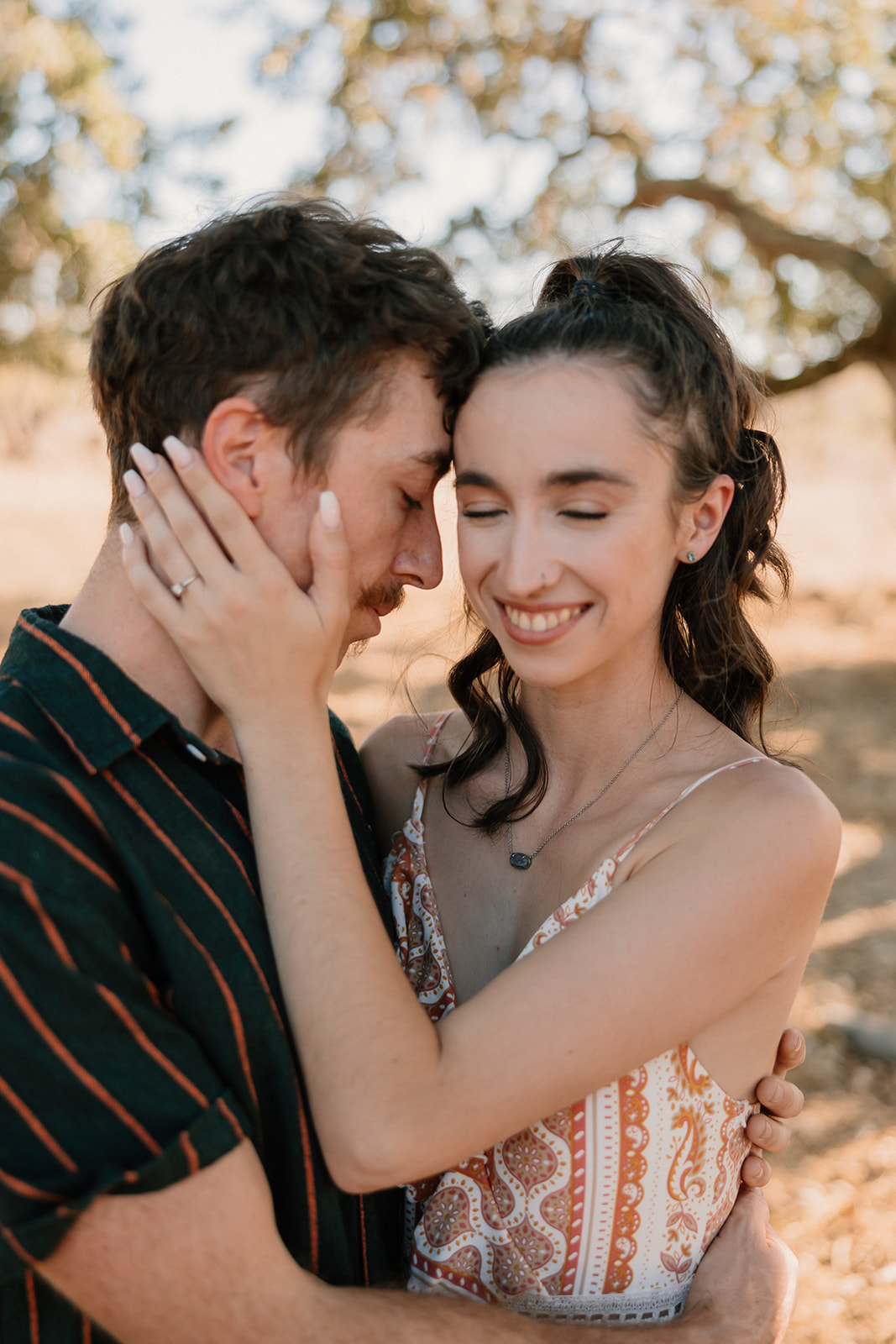 A couple embracing under a tree, exchanging a loving gaze, dressed in casual summer attire for their park engagement photos.