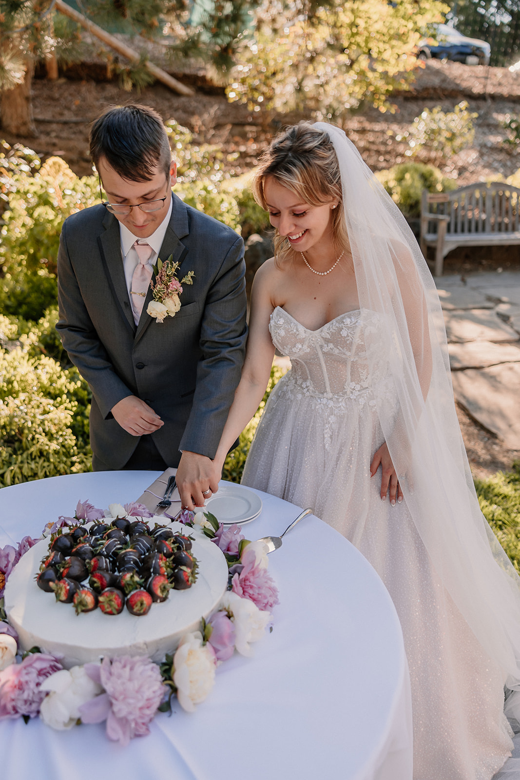 A groom in a gray suit is feeding cake to a bride in a white wedding dress and veil. They are standing outdoors near a table with flowers and cake.
