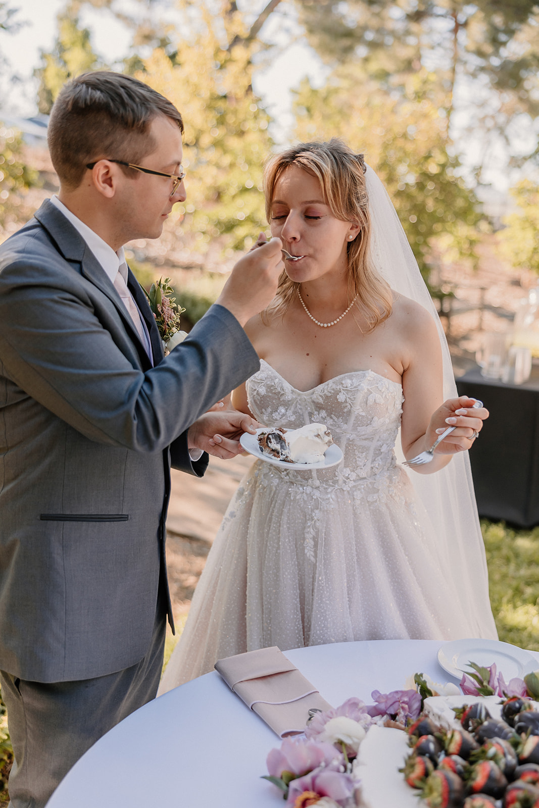 A groom in a gray suit is feeding cake to a bride in a white wedding dress and veil. They are standing outdoors near a table with flowers and cake.