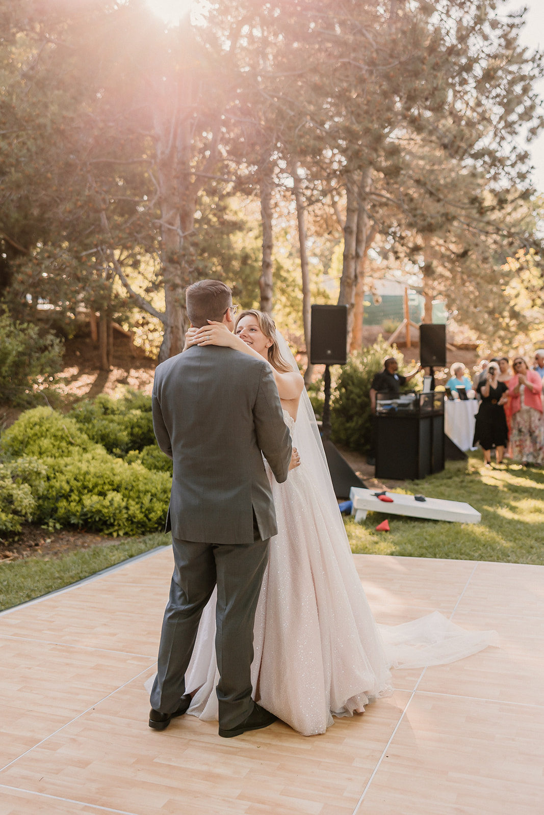 A couple, wearing a suit and a gown, share a kiss on an outdoor dance floor surrounded by trees and greenery, under the sunlight at their wedding at the gardens in heather farms