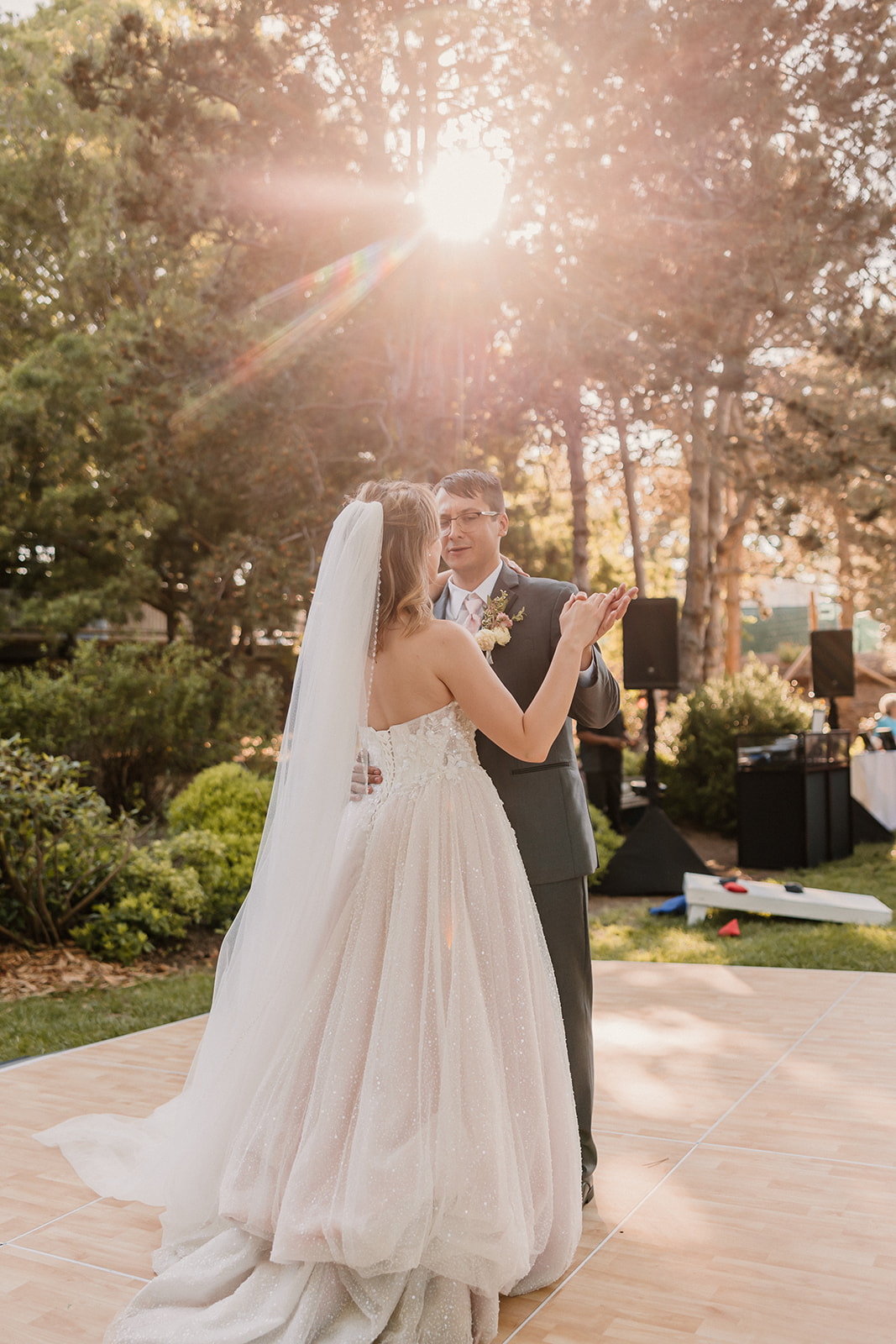 A couple, wearing a suit and a gown, share a kiss on an outdoor dance floor surrounded by trees and greenery, under the sunlight at their wedding at the gardens in heather farms