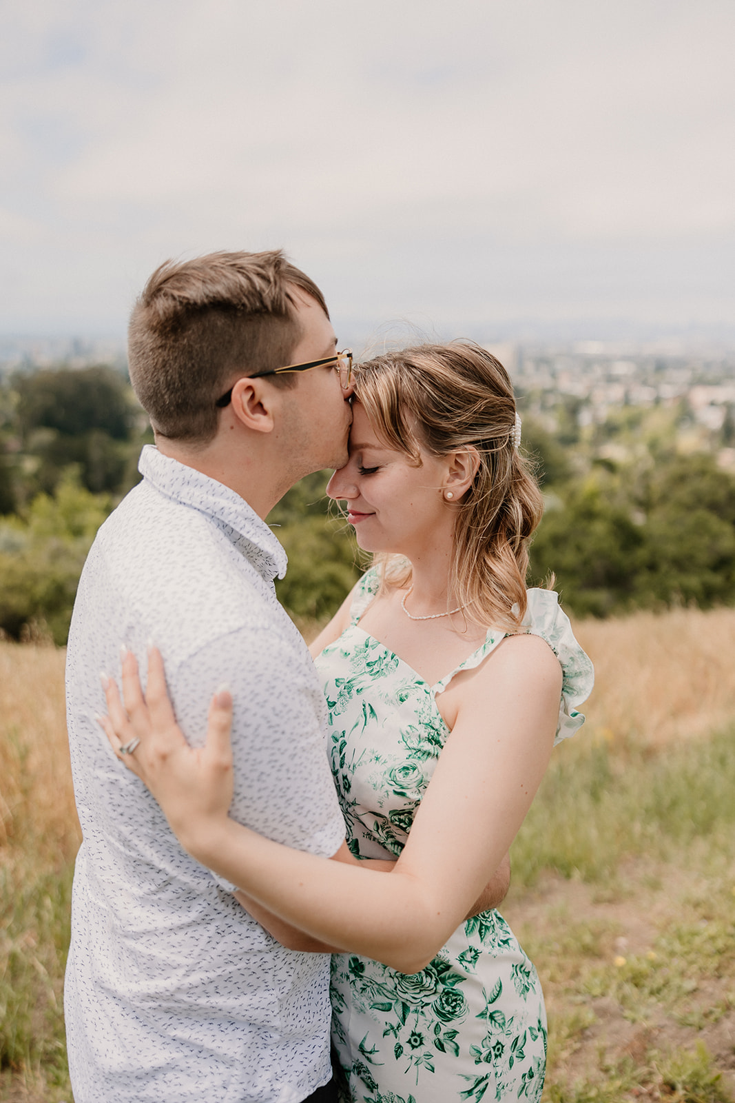 A young couple stands closely with foreheads touching, embracing each other in a grassy outdoor setting under a partly cloudy sky at their day after session