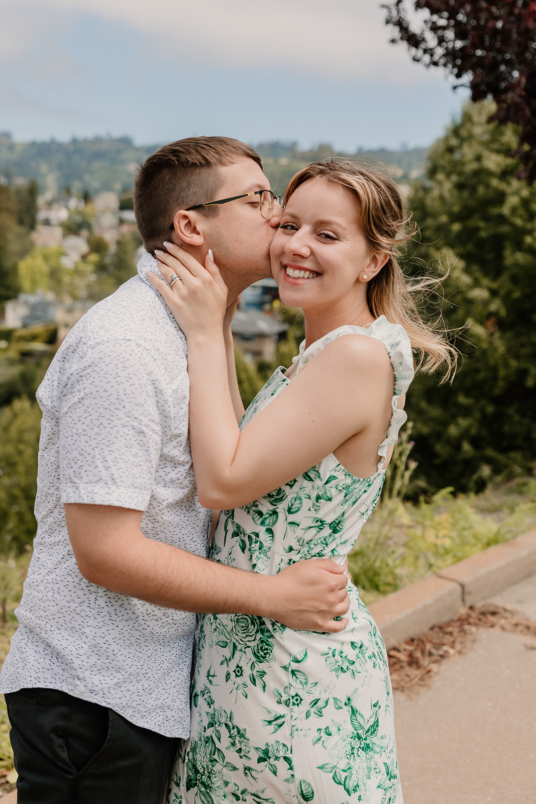 A young couple stands closely with foreheads touching, embracing each other in a grassy outdoor setting under a partly cloudy sky at their day after session