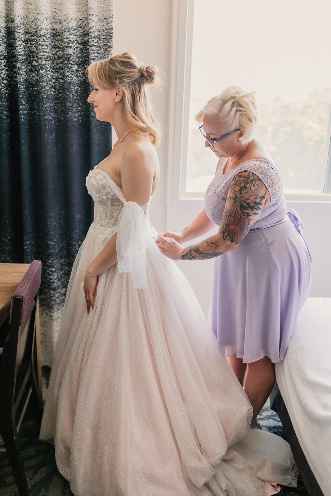 A woman in a wedding dress stands as another woman in a lavender dress adjusts the back of the gown.