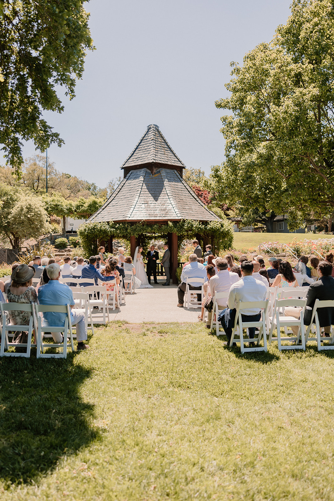 A bride and groom stand facing each other during an outdoor wedding ceremony. The bride wears an off-shoulder gown with a long train, and the groom wears a gray suit. They are surrounded by greenery at their wedding at the gardens at heather farms