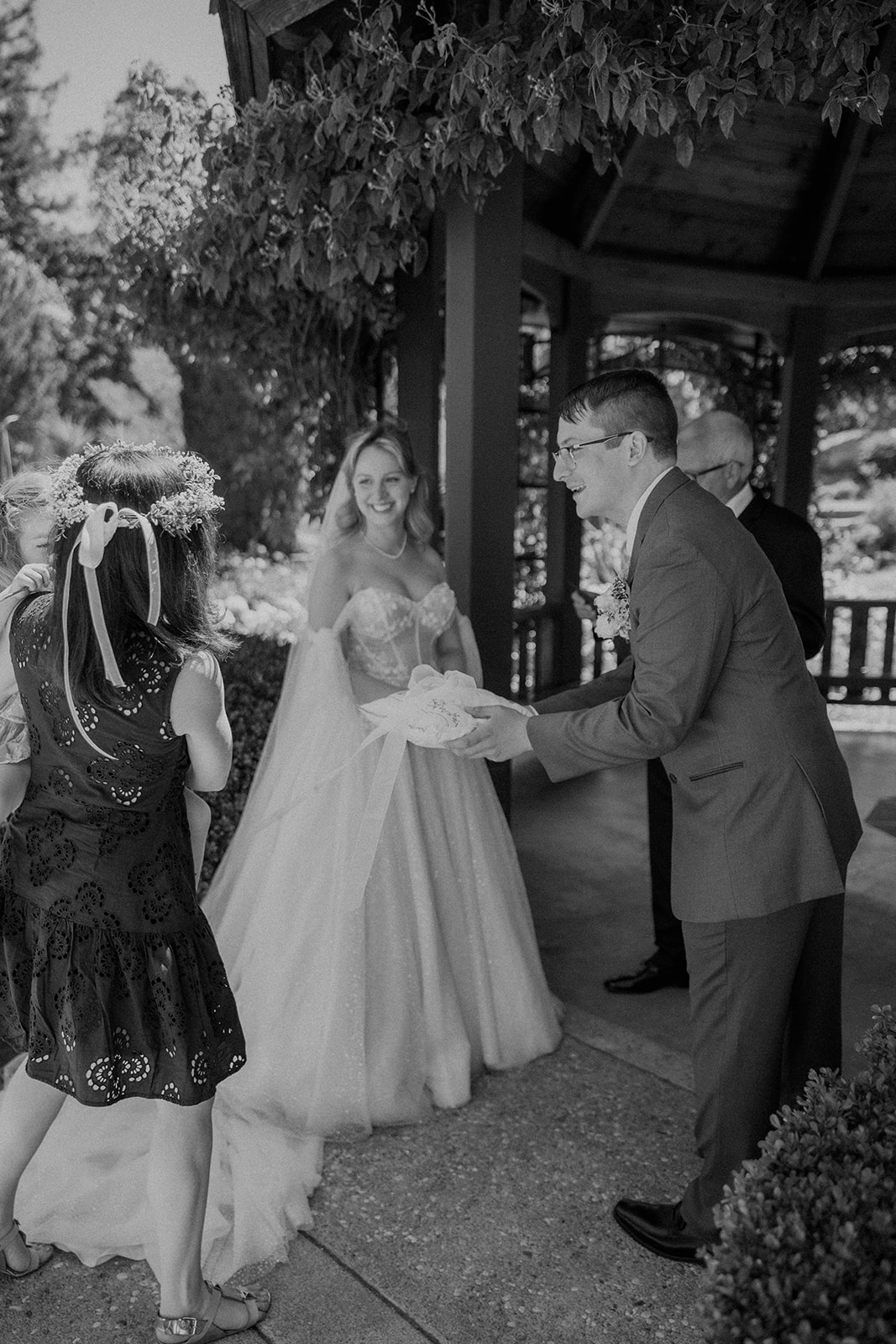 A bride and groom stand facing each other during an outdoor wedding ceremony. The bride wears an off-shoulder gown with a long train, and the groom wears a gray suit. They are surrounded by greenery at their wedding at the gardens at heather farms