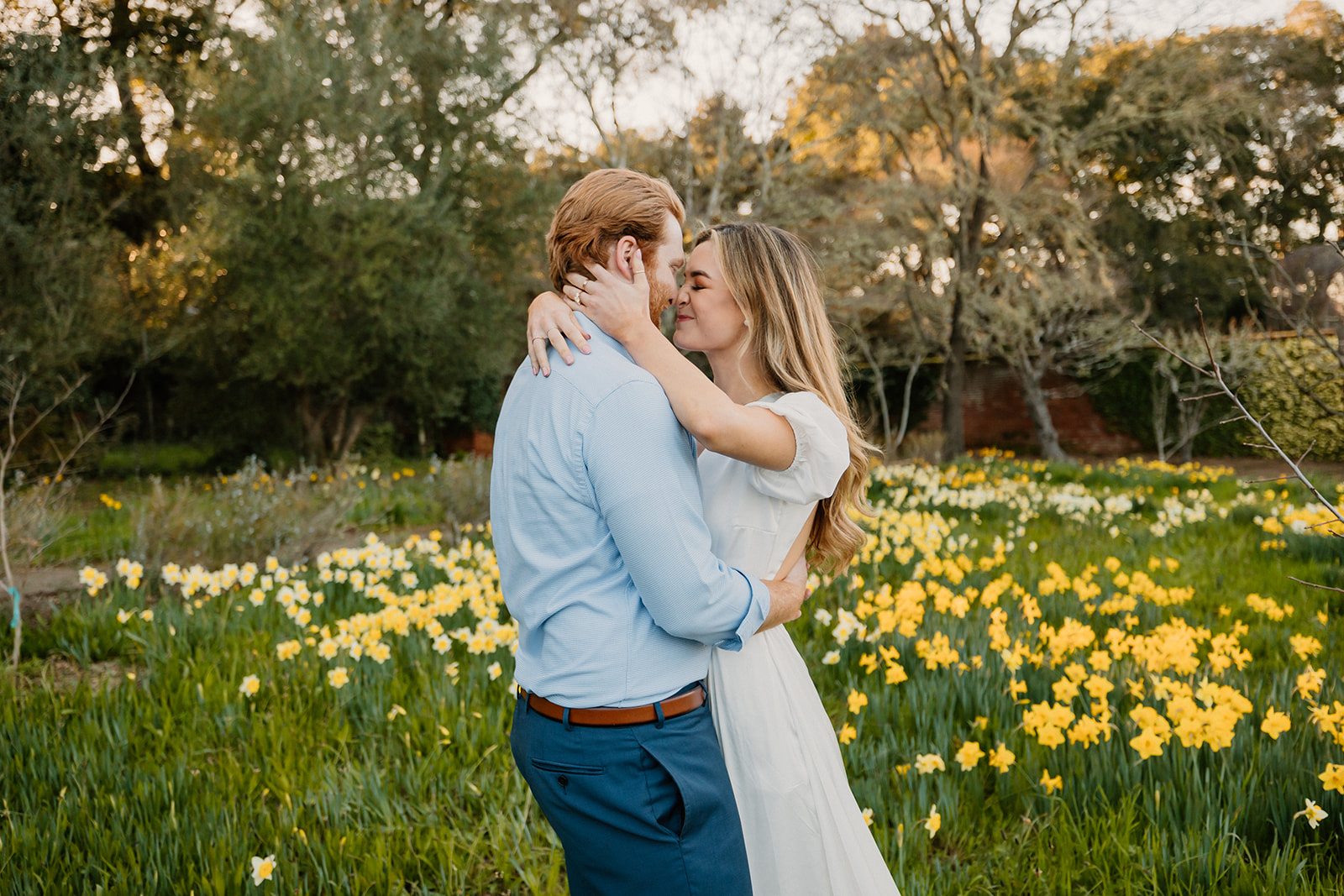 A couple embracing and smiling at, taking their engagement photos, both dressed in casual attire.