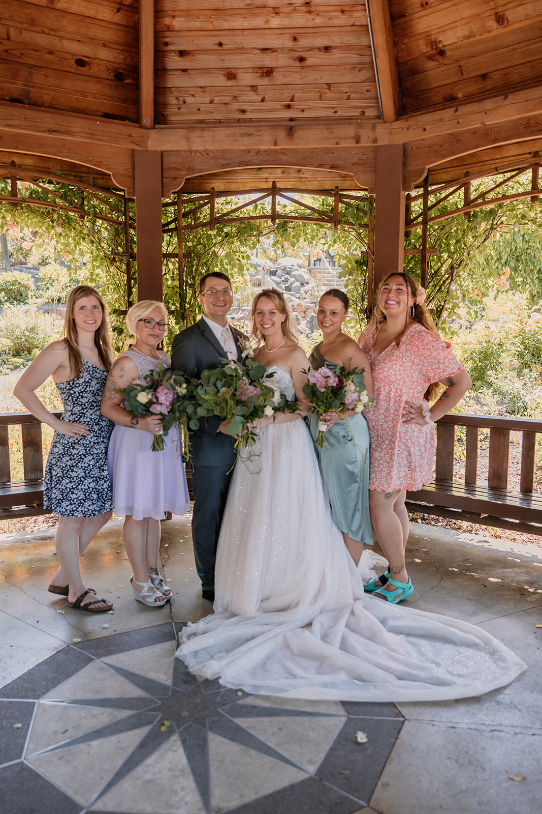 A bride and groom stand together under a gazebo, with women posing with them. The bride holds a bouquet, and all are smiling.