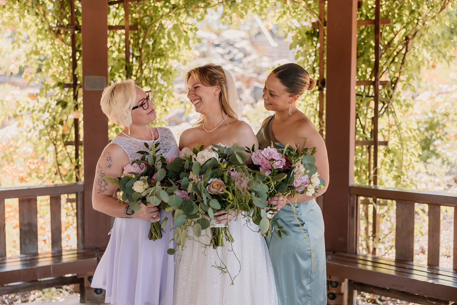 A bride and groom stand together under a gazebo, with women posing with them. The bride holds a bouquet, and all are smiling.
