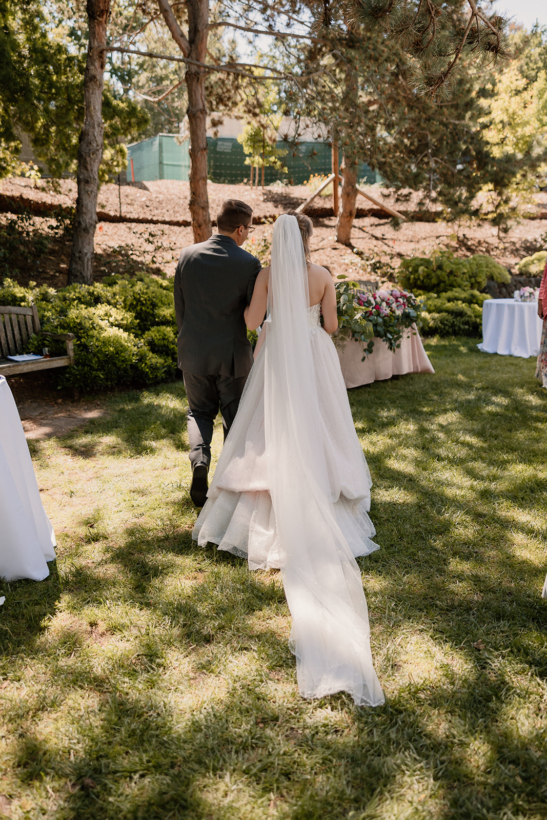 A bride and groom smiling and walking together outdoors through an open gate, surrounded by greenery and sunlight.