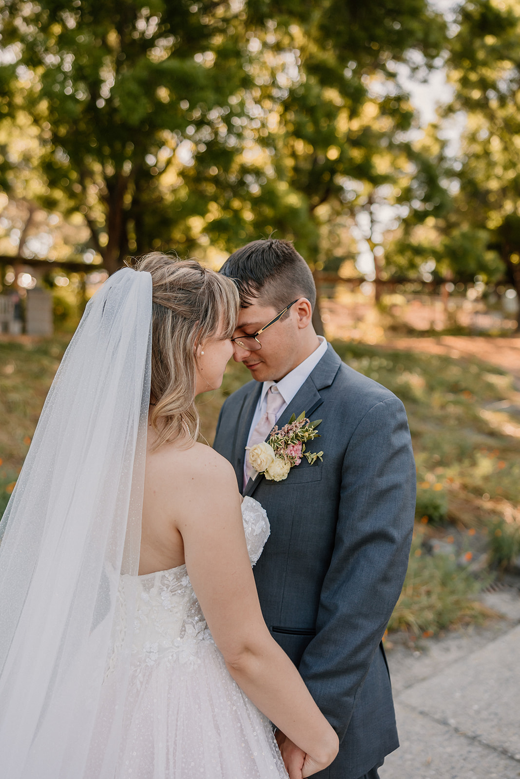 A bride and groom share a kiss outdoors in a garden setting, with the bride holding a large bouquet of flowers and a scenic background of trees and a pond at their wedding at the gardens at heather farms