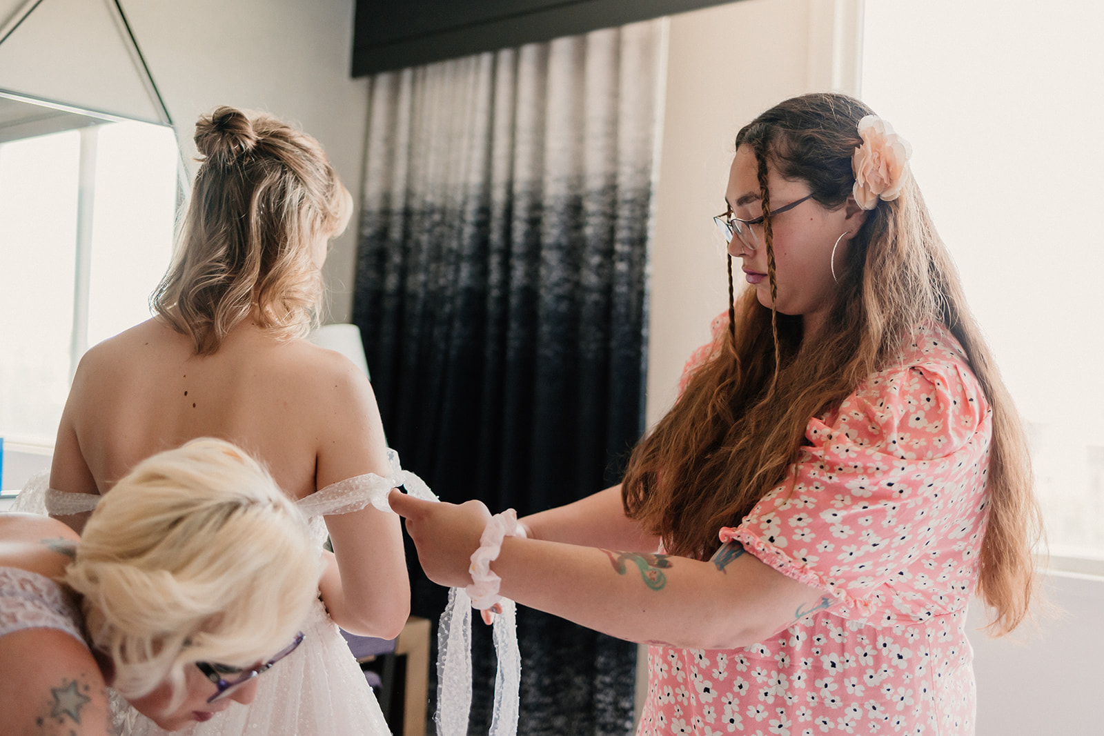 A woman in a wedding dress stands as another woman in a lavender dress adjusts the back of the gown.