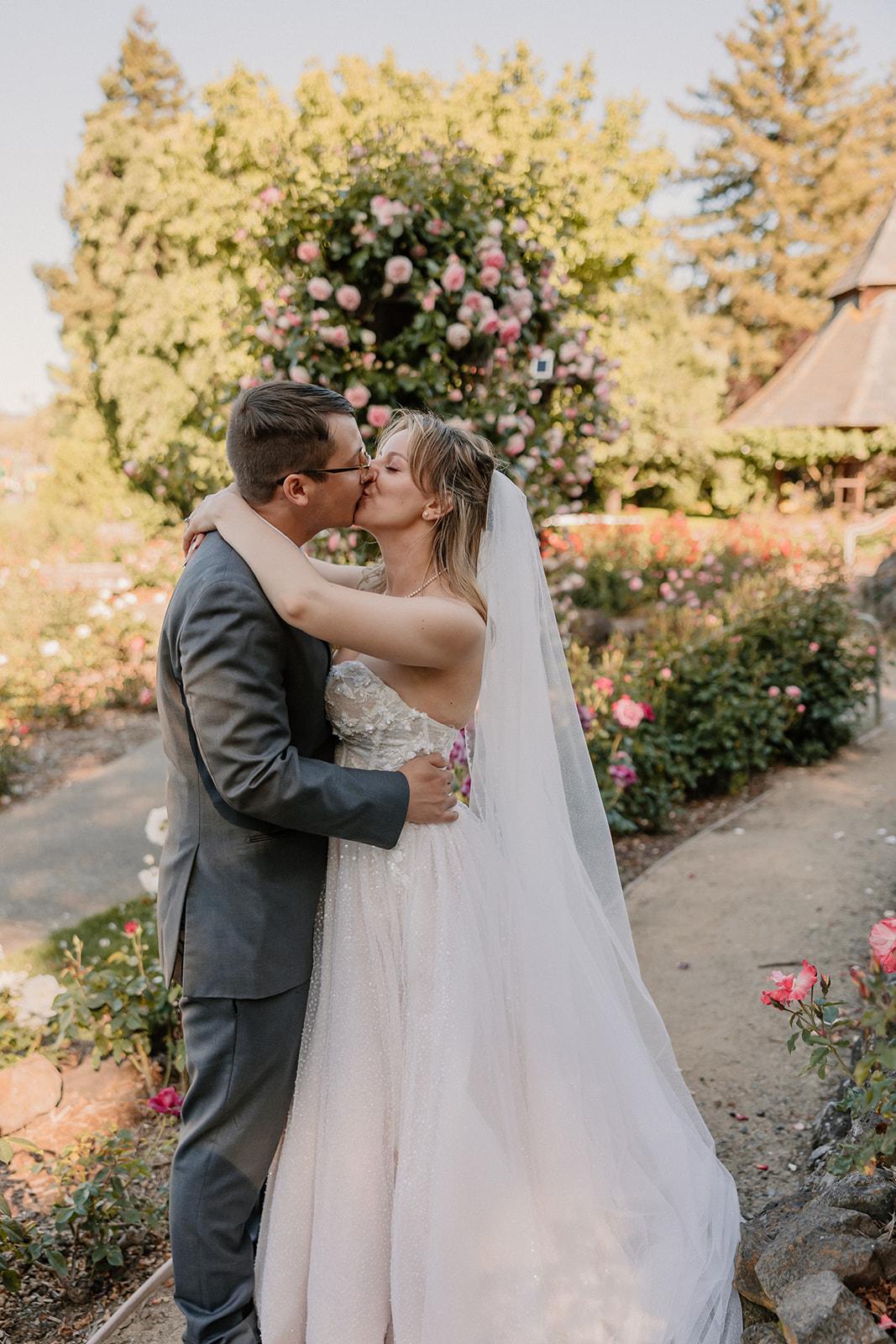 A bride and groom share a kiss outdoors in a garden setting, with the bride holding a large bouquet of flowers and a scenic background of trees and a pond at their wedding at the gardens at heather farms