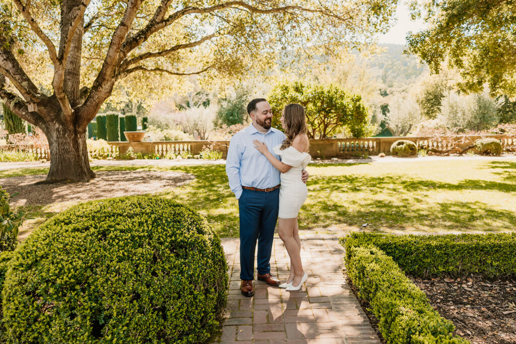 A man and woman stand together, smiling, in a sunny garden with well-maintained hedges and trees in the background. The woman is dressed in white and the man is dressed in bluefor their engagement photos at the filoli gardens