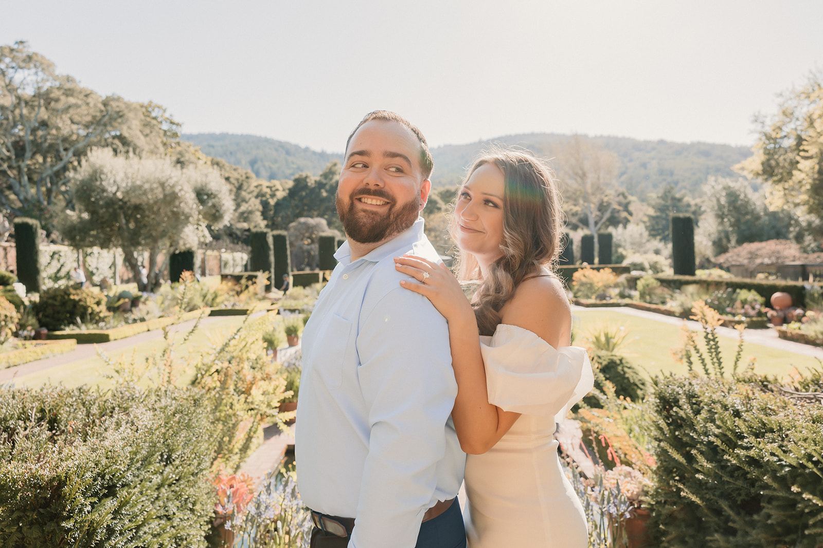 A man in a blue shirt kisses a woman in a white dress on the cheek while standing in a sunlit garden for their engagement photos at the filoli gardens