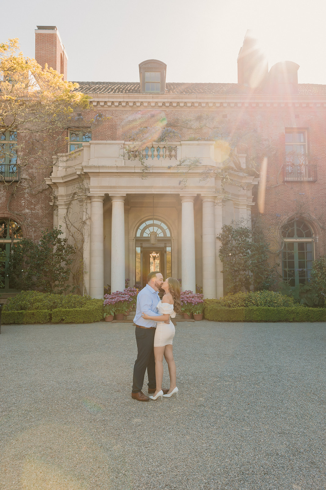 A couple embraces and kisses in front of a large historic building with sunlight streaming in from the top right corner.