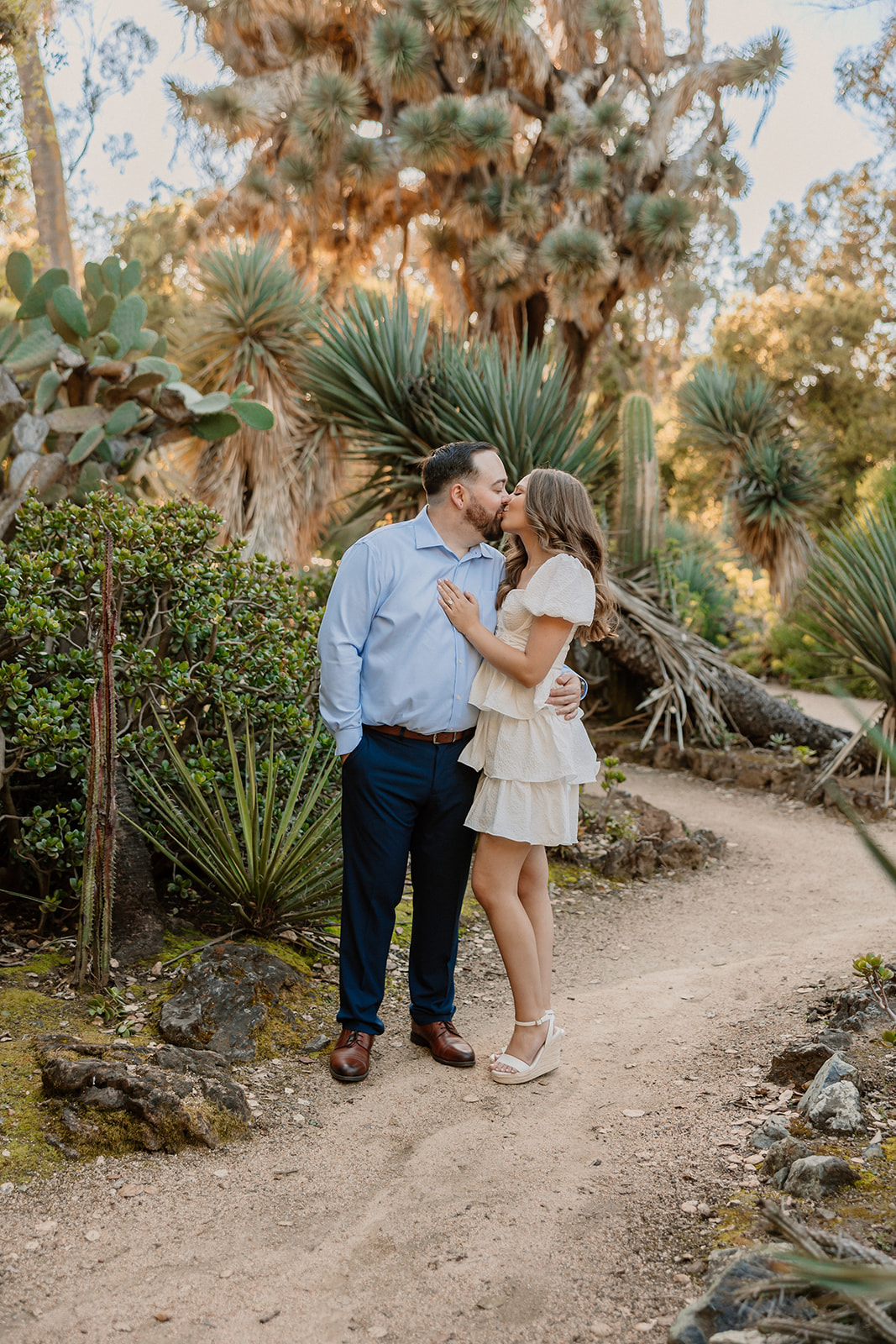 A man and woman stand on a dirt path in a garden, sharing a kiss. The man is in a blue shirt and dark pants, and the woman is in a white dress. Greenery and various plants surround them for their engagement photos at cactus garden in stanford