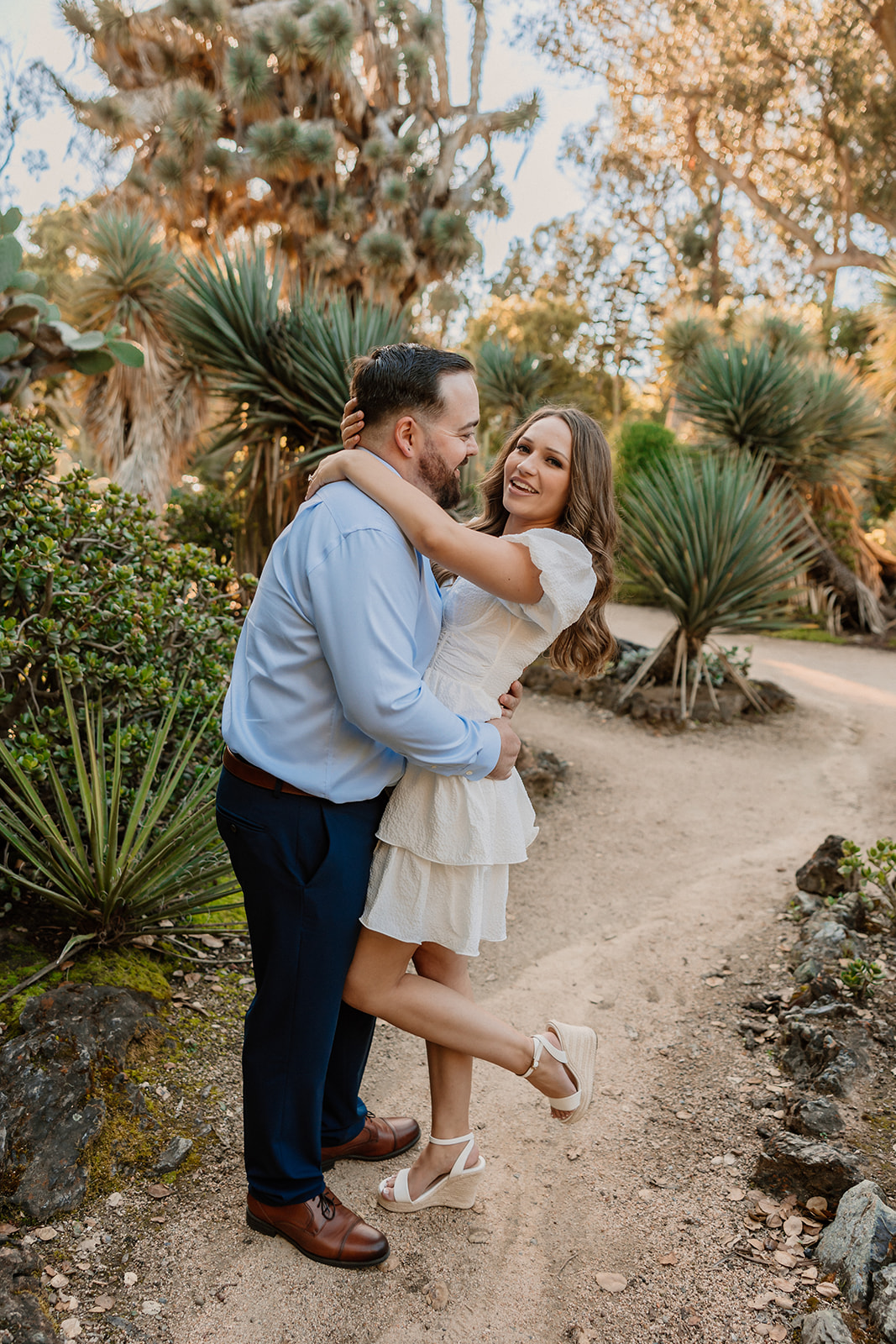 A man and woman stand on a dirt path in a garden, sharing a kiss. The man is in a blue shirt and dark pants, and the woman is in a white dress. Greenery and various plants surround them for their engagement photos at cactus garden in stanford