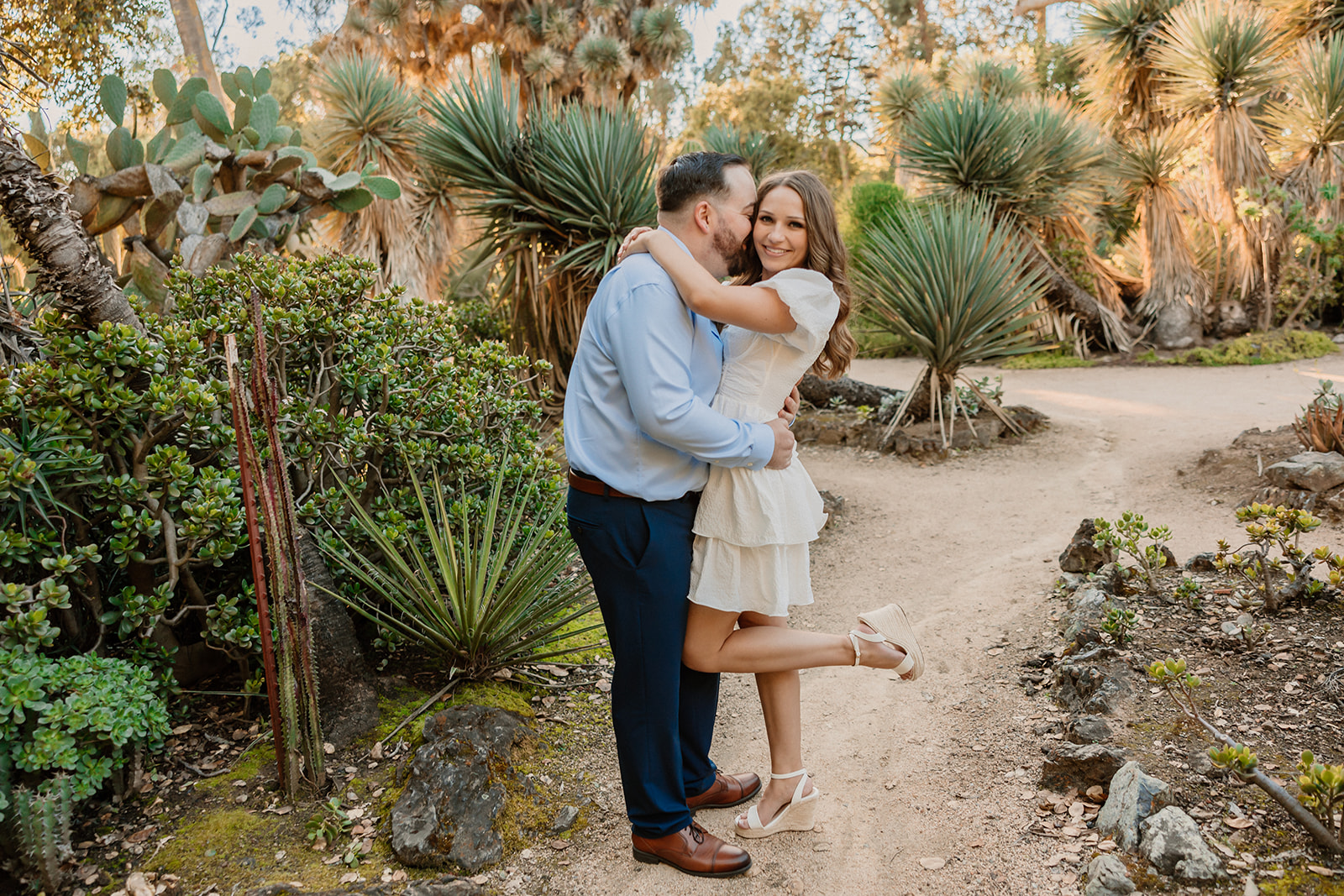 A couple embraces on a dirt path surrounded by lush greenery and various plants. The woman wears a white dress and lifts one leg, while the man wears a blue shirt and dark pants at their engagement photos at cactus garden in stanford