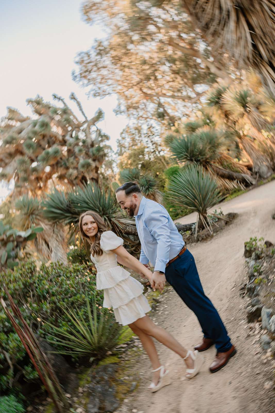 A couple embraces on a dirt path surrounded by lush greenery and various plants. The woman wears a white dress and lifts one leg, while the man wears a blue shirt and dark pants  at cactus garden in stanford