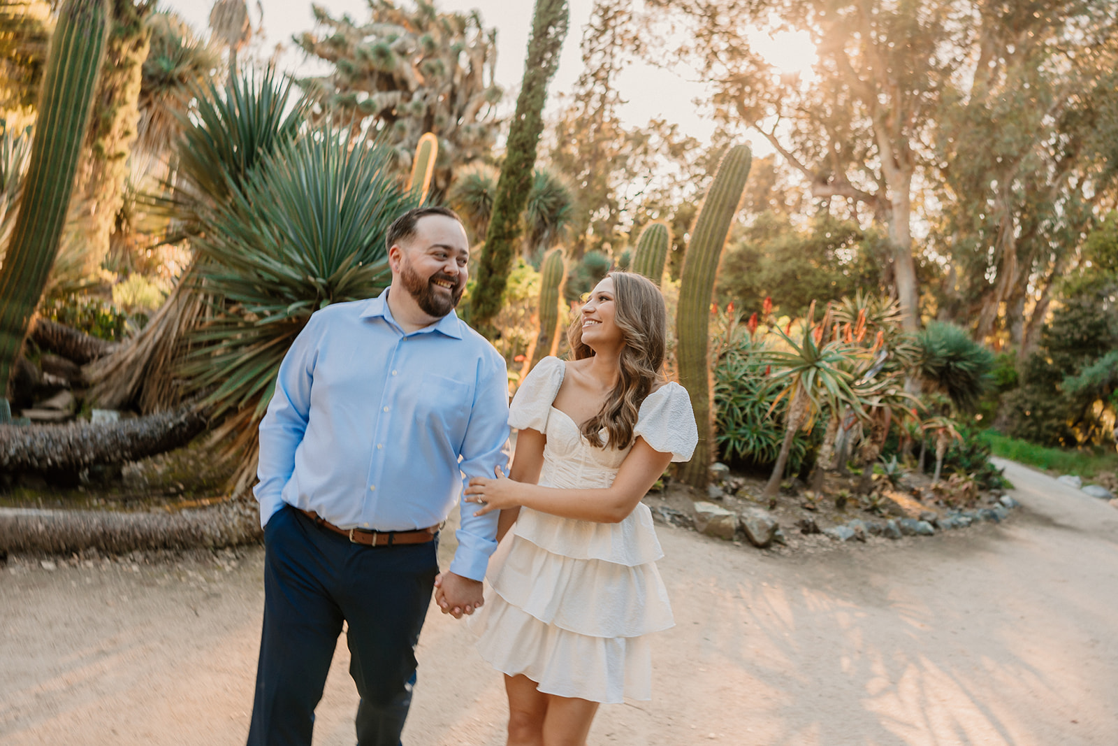 A couple embraces on a dirt path surrounded by lush greenery and various plants. The woman wears a white dress and lifts one leg, while the man wears a blue shirt and dark pants at cactus garden in stanford