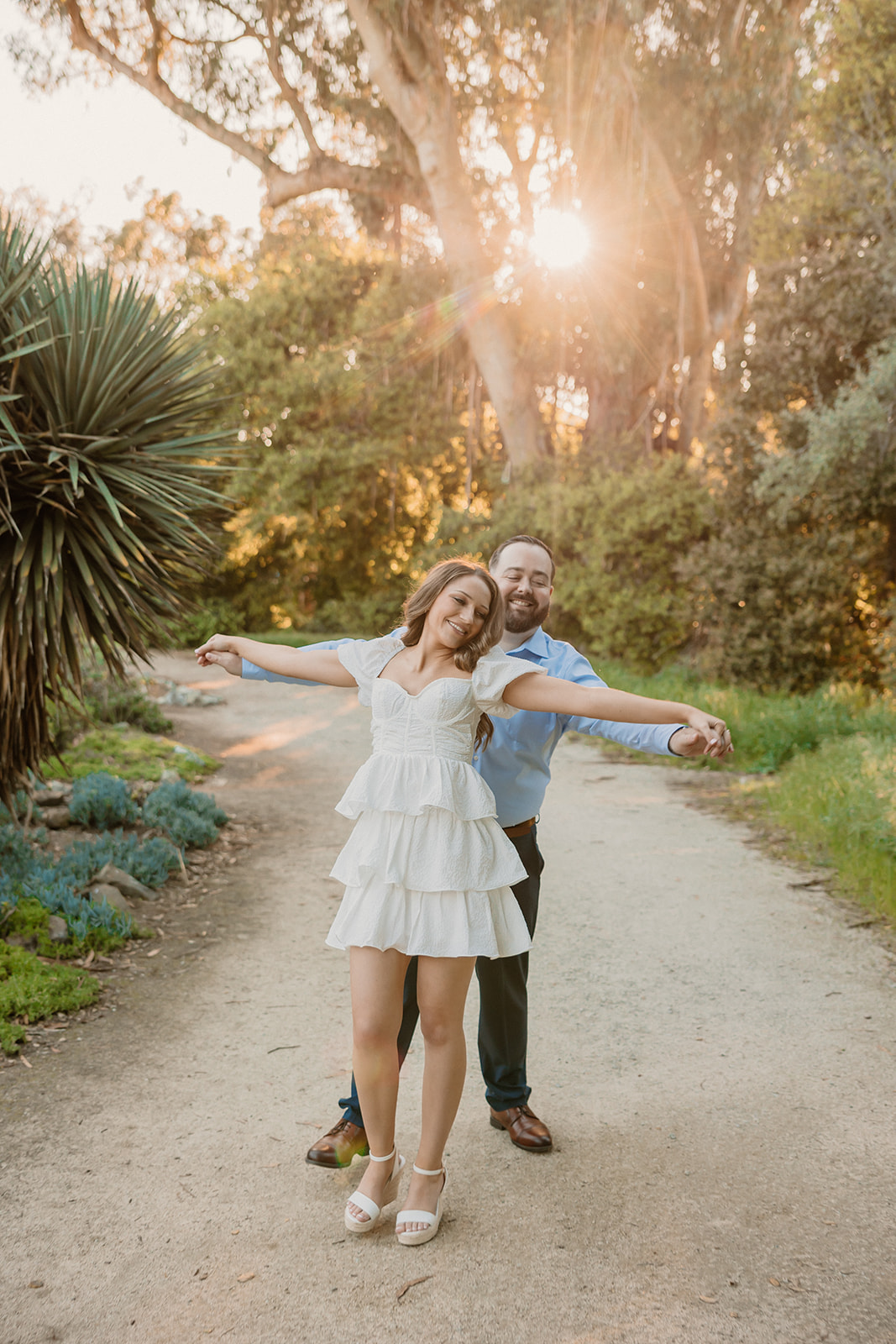 A couple embraces on a dirt path surrounded by lush greenery and various plants. The woman wears a white dress and lifts one leg, while the man wears a blue shirt and dark pants at cactus garden in stanford