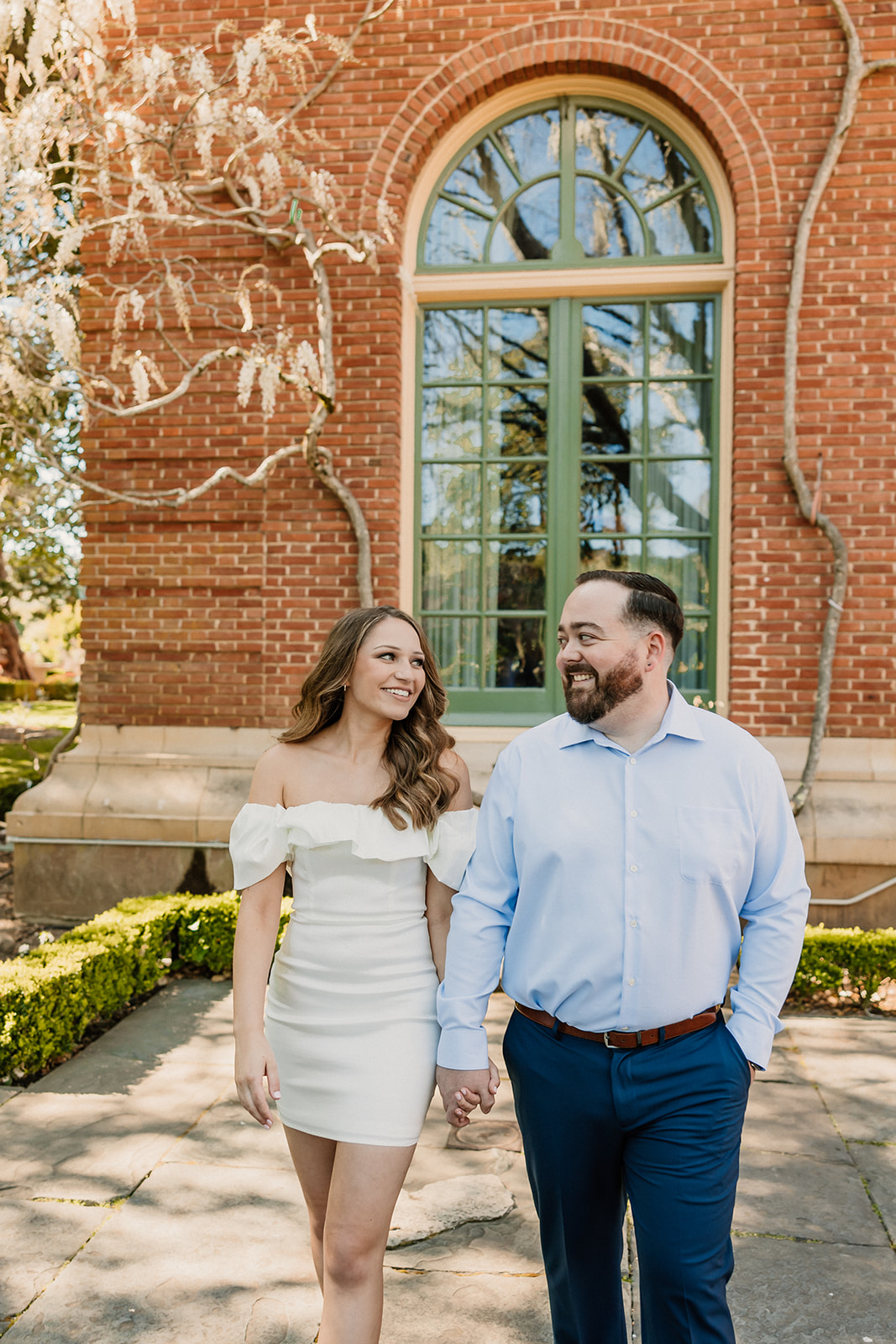 A couple stands hand in hand in front of a large arched window on a brick building, surrounded by greenery and vines for their engagement photos at filoli gardens